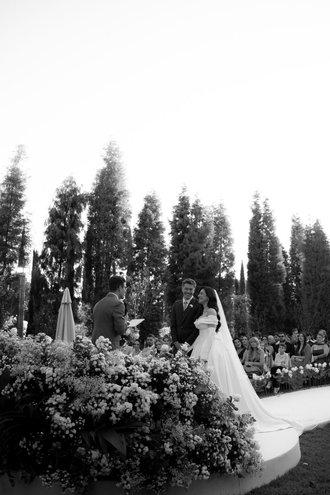 Murilo Mestriner celebrando um casamento ao ar livre sob um arco de flores, em preto e branco